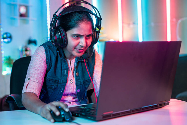 Smiling girl playing live video game on laptop by talking on headphones at home on neon background - concept of vlogger, gaming addiction and championship. - Foto, immagini