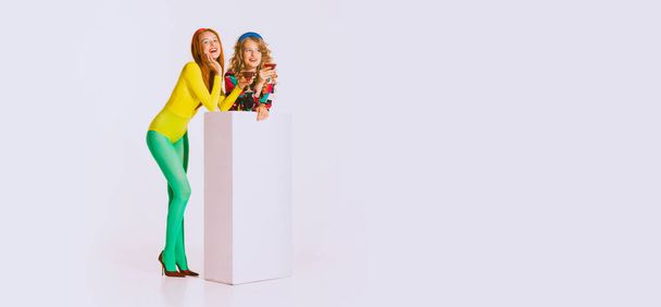 Portrait of two stylish, beautiful, young girls in bright, colorful retro clothes standing, talking isolated over grey background. Party. Concept of youth, retro style, 90s era, fashion, lifestyle, ad - Photo, image