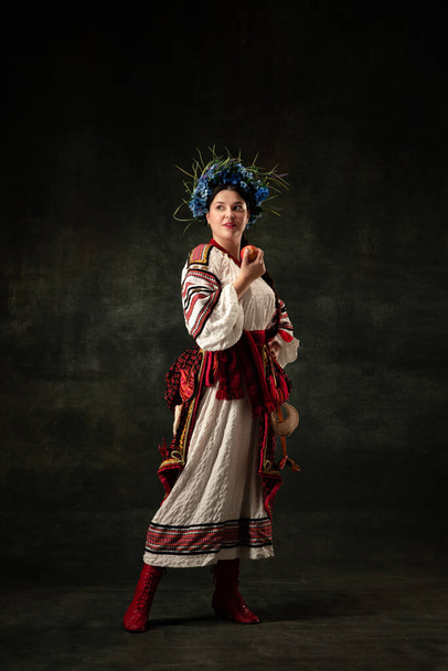 Freedom and independence. Art portrait of beautiful woman wearing traditional folk Ukrainian costume posing isolated over dark vintage background. Fashion, beauty, cultural heritage concept. - 写真・画像