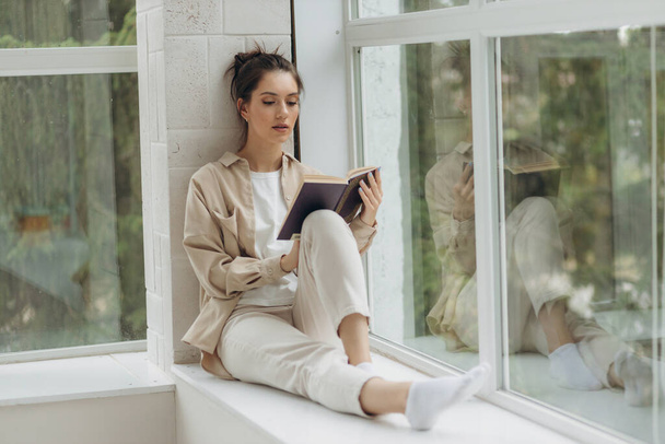 Красива молода жінка сидить біля вікна і читає книгу
. - Фото, зображення