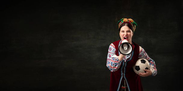 Flyer with expressive young woman in national traditional folk Ukrainian attire shouting at megaphone isolated over dark vintage background. Sport, freedom, rights, fashion, beauty, betting concept. - Foto, imagen