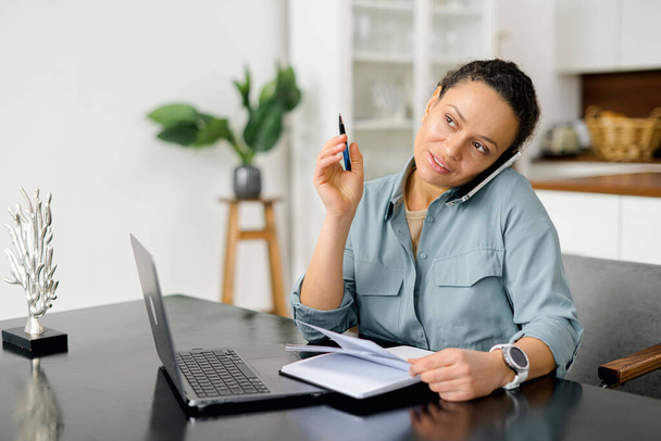 Freelance woman talks on the smartphone and takes notes with pen, using laptop for remote work from home, write down some information sitting at the table. Multitasking concept - Photo, Image