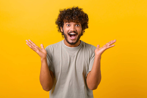 Astonished Indian young curly man screams wow and looking at the camera with mouth wide opened, surprised happy guy excited with incredible promotion, standing isolated on yellow - Фото, зображення