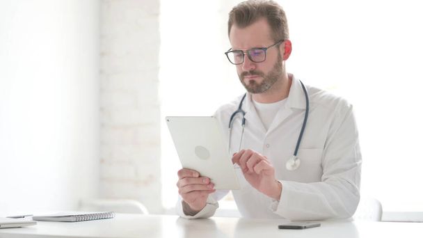 Middle Aged Doctor using Tablet while Sitting in Clinic - Foto, Imagen
