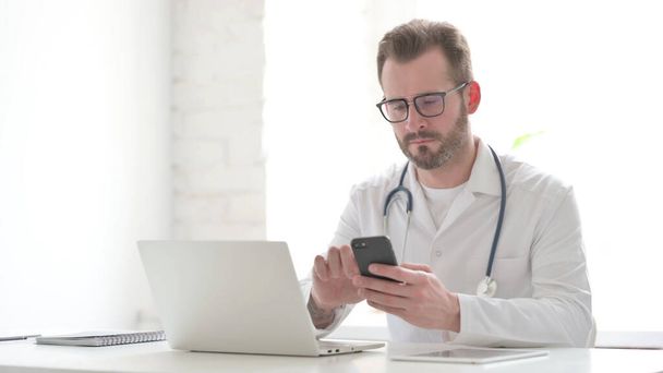 Middle Aged Doctor using Smartphone while using Laptop in Clinic - Foto, immagini