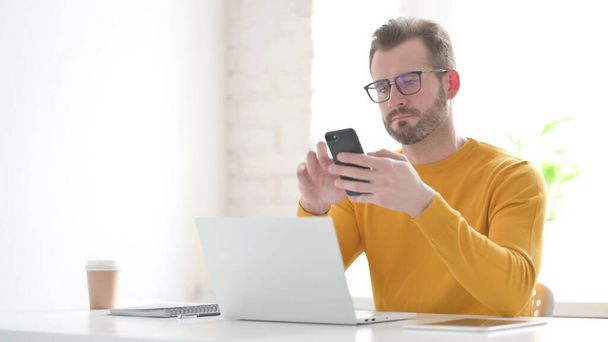 Középkorú férfi használ okostelefon használata közben Laptop az irodában - Fotó, kép