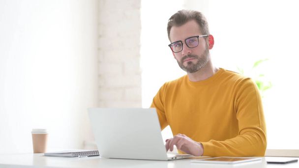 Hombre de mediana edad mirando a la cámara mientras usa el ordenador portátil en la oficina - Foto, imagen