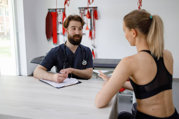Doctor physiotherapist and young fitness woman make a training plan for rehabilitation. Dialogue between a sports doctor and an athletic girl in the office of a health center - Zdjęcie, obraz