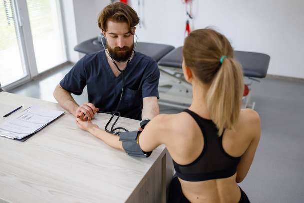 Doctor physiotherapist measuring pressure to a young fitness woman sitting at the rehabilitation gym - Foto, immagini