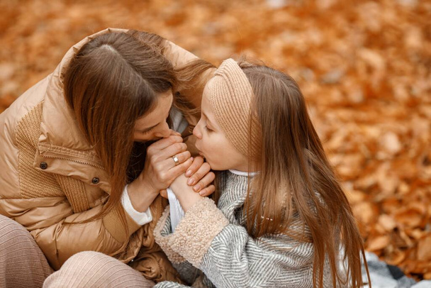 Young woman and little girl in autumn forest. Woman and her daughter sitting on a fallen leaves and holding hands. Girl wearing fashion grey jacket and woman brown one. - Foto, afbeelding