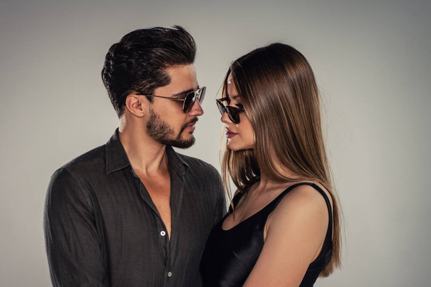 Portrait of young handsome couple wearing fashionable sunglasses, looking at eachother. Studio background. Bearded man posing with beautiful woman. - Fotografie, Obrázek
