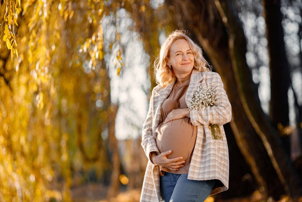 Portrait of middle-aged pregnant woman outdoors at park. Middle age pregnant woman expecting baby at aged pregnancy. Blonde woman wearing brown sweater and beige coat. - Foto, afbeelding