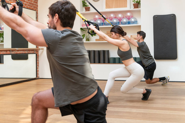 Спортсмени і спортсмени в активному одязі роблять TRX перетинаючи баланс лаунж вправи разом під час тренування підвіски в тренажерному залі
 - Фото, зображення