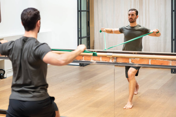 Back view of concentrated young bearded male athlete in sportswear pulling resistance band while doing shoulder stability exercise in front of mirror in modern fitness center - Zdjęcie, obraz