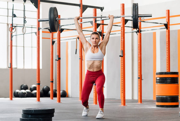Full body strained sportswoman lifting heavy barbell over head and lunging while doing split jerk exercise during intense weightlifting training in gym - Photo, Image