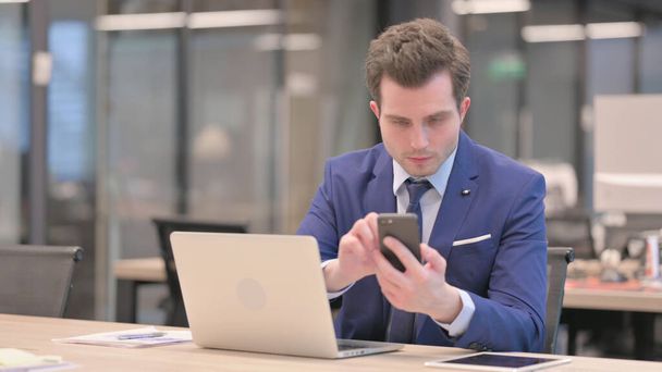 Geschäftsmann mittleren Alters benutzt Smartphone, während er Laptop im Büro benutzt - Foto, Bild