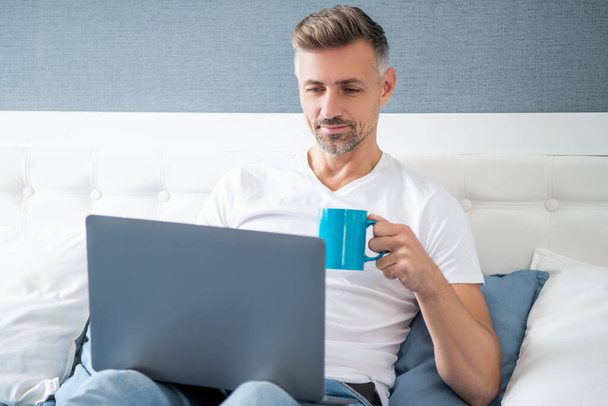 smiling mature man using computer in bed with coffee. - Foto, immagini