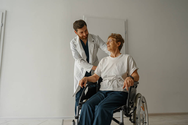 Профессиональный врач-мужчина несет пациента на инвалидной коляске в медицинском зале клиники. Высокое качество фото - Фото, изображение