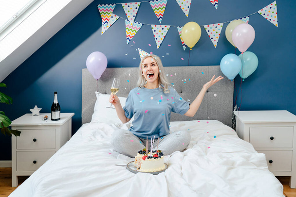 Happy emotional woman in pajama and party cap with champagne glass throwing confetti up while sitting on the bed in a decorated bedroom.Time for yourself Celebration at home. Happy birthday concept. - Фото, изображение