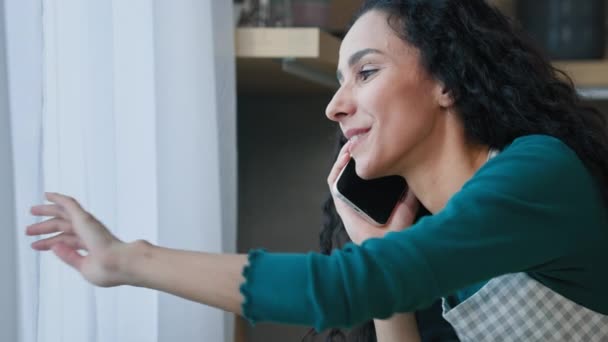 Smiling young arabian woman chatting with friend by cellphone has pleasure phone conversation gossip stand in house look at window greet friend from outside quarantine self-isolation due covid concept - Materiał filmowy, wideo