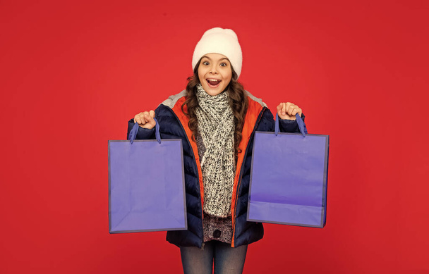 surprised kid in puffer jacket and hat. teen girl after shopping on red background. child holding shopping bags with copy space. winter sales. - 写真・画像