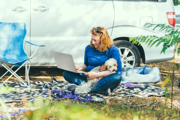 Adult woman working on laptop, sitting with her cockapoo puppy on the background of camper car recreational vehicle. Female living on a rv motorhome with animals and travel the world. Best friend. - Fotografie, Obrázek