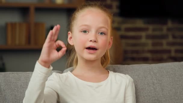 Portrait of caucasian happy child girl smiling kid sitting on sofa at room looking at camera showing ok gesture everything fine like sign positive good emotion. Little cute charming teen kid recommend - Imágenes, Vídeo