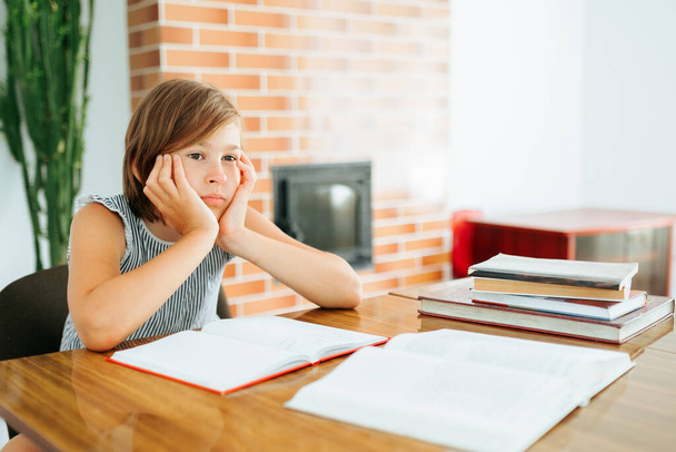 Front view blurred background beautiful pensive girl kid sitting elbows on table hands holding head. Child is sad sits on chair over notebooks and dreams about something. Books on the table. - Фото, зображення