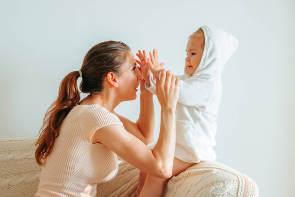 Side view young mum with baby child sits on sofa mother hold hands of kid play hide and seek with palms. Mother cover face with daughter hands. Background a bright living room, white walls.  - Fotó, kép