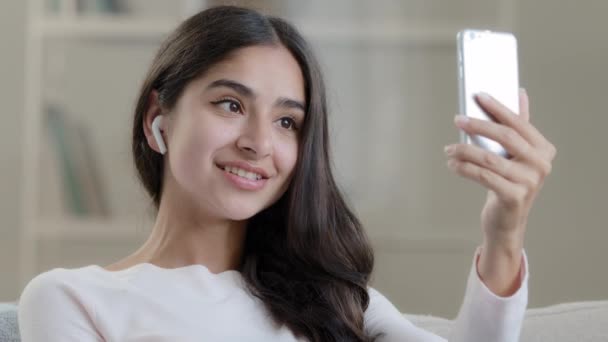 Female blogger arabic young girl beautiful pretty woman influencer in wireless earphones headphones talks at mobile camera takes self photo streaming live video chat records video blog talks distance - Πλάνα, βίντεο