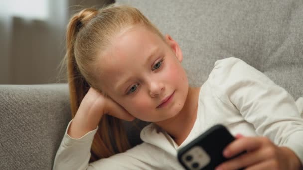 Relaxed blonde child little girl blogger cute kid teen resting on couch playing online games on mobile phone posting photos in social media apps chatting with friends watching funny cartoons at home - Záběry, video