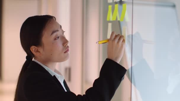 Brainstorming process. Close up portrait of pensive asian woman manager writing on sticky notes, planning project work near glass wall, slow motion, free space - Imágenes, Vídeo