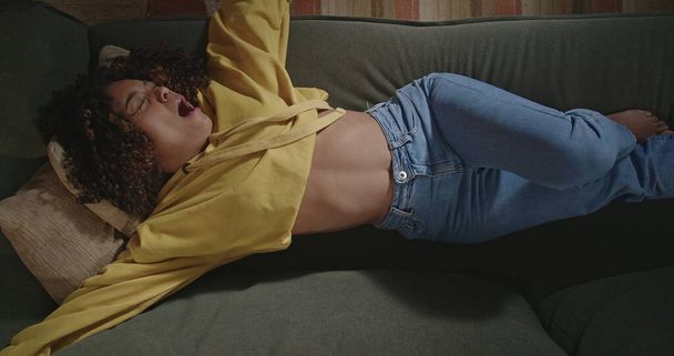 Young woman stretching body resting on sofa. From above angle of black girl relaxing on couch in evening night checking her phone. Person using smartphone - Фото, изображение