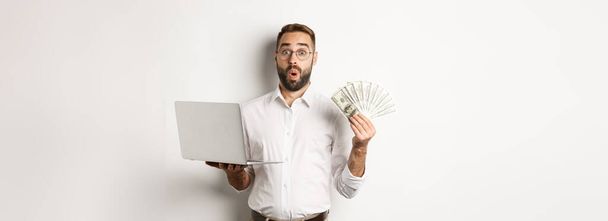 Zaken en elektronische handel. Man kijkt verbaasd met geld inkomen, werken online, met behulp van laptop, staande over witte achtergrond. - Foto, afbeelding