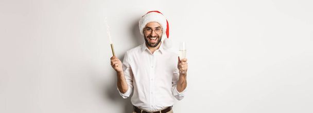 Winter holidays and celebration. Handsome bearded man having New Year party, holding firework sparkle and champagne, wearing santa hat, white background. - Foto, Bild