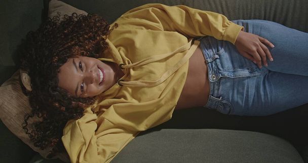 Young brunette woman in yellow blouse smiles at camera while resting on her sofa in living room. One happy African American girl. From above angle of a Brazilian black girl - 写真・画像
