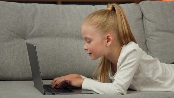 Carefree little kid pretty blonde girl child daughter using laptop lying on grey sofa surfing internet chatting with classmates playing games studying online at home. Children tech addiction concept - 映像、動画