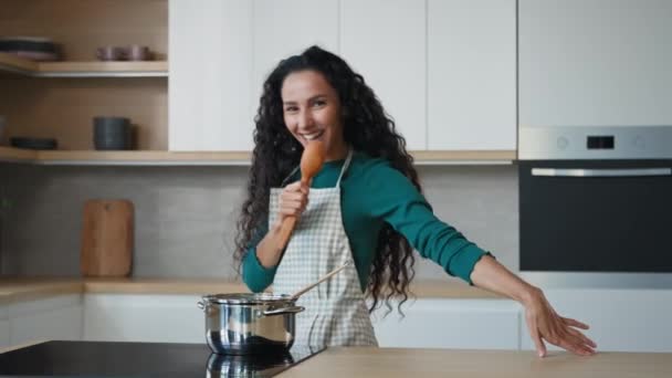 Active playful arabian woman singer mistress with long curly hair rhythmic moves to music singing popular song in culinary wooden spoon cooking equipment flatware having fun domestic karaoke at home - Materiaali, video