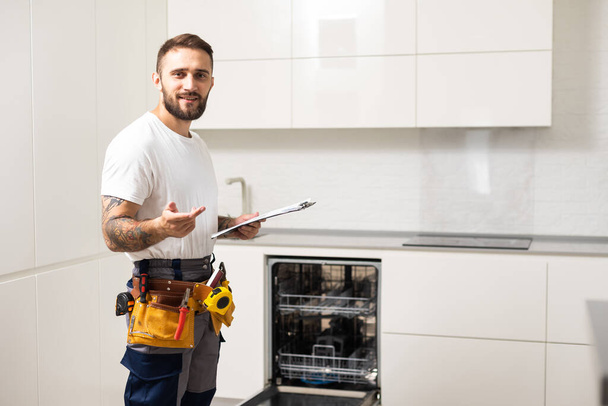 Ev mutfağındaki bulaşık makinesinin tamiri sırasında işçi kıyafeti giymiş genç modern bir tamirci.. - Fotoğraf, Görsel