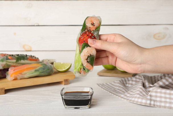 Woman dipping roll wrapped in rice paper into sauce at white wooden table, closeup - 写真・画像