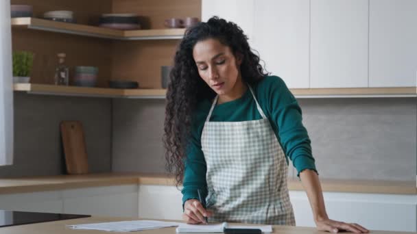 Arabian hispanic housewife pretty curly woman wears apron stand in kitchen write notes food recipe use calculator for counting calories checklist of products calculate domestic bills paperwork at home - Záběry, video