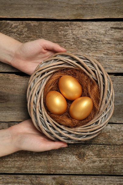 Vrouw houdt nest met glanzende gouden eieren aan houten tafel, bovenaanzicht - Foto, afbeelding