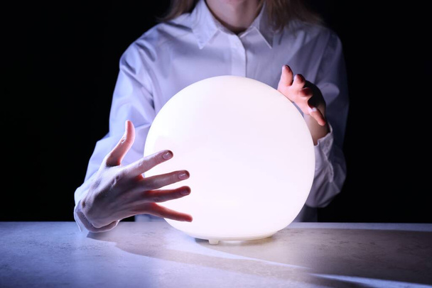 Businesswoman using glowing crystal ball to predict future at table in darkness, closeup. Fortune telling - Fotografie, Obrázek