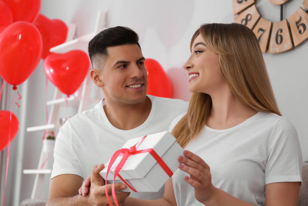 Man presenting gift to his girlfriend in room decorated with heart shaped balloons. Valentine's day celebration - Φωτογραφία, εικόνα
