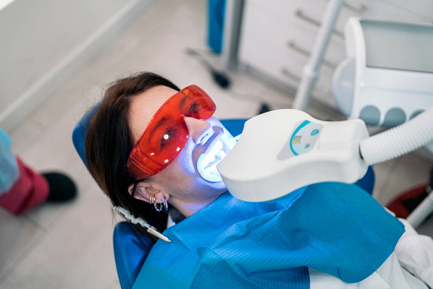 Woman lying down in dental chair and wearing protective glasses using tooth whitening machine. - Fotoğraf, Görsel