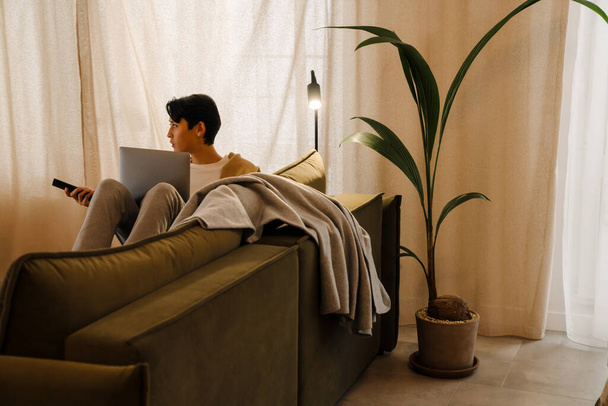 Asian boy watching tv while working with laptop on sofa at home - Foto, imagen