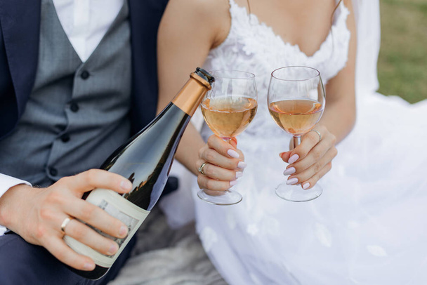 Close up cropped unrecognizable groom and bride hands drinking and pouring bubbly champagne wine from bottle and glasses together, sitting on blanket in park and having fun on wedding celebration day. - Photo, image