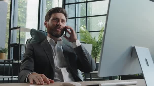 Busy millennial bearded man in office company CEO worker manager consulting client by telephone sitting at table with computer. Professional broker salesman remote mobile phone conversation ecommerce - Záběry, video