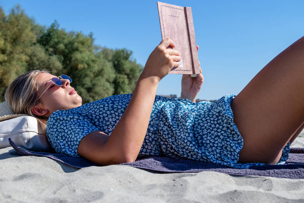 A beautiful young woman with sunglasses reading a book, and enjoying her time alone on a deserted sandy beach.  - Φωτογραφία, εικόνα