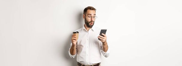 Empresario bebiendo café y mirando sorprendido en el mensaje en el teléfono móvil, de pie sorprendido sobre fondo blanco. - Foto, imagen